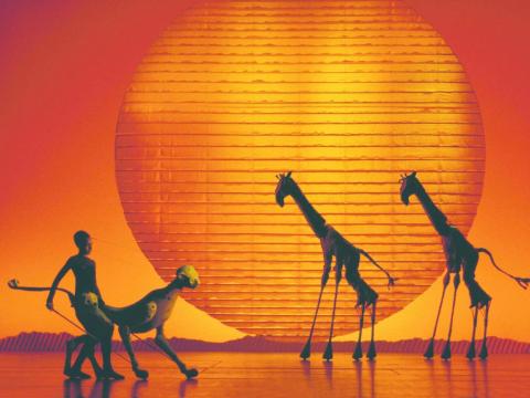 Production image of Disney's The Lion King with safari animal puppets in front of a large orange sunset background.